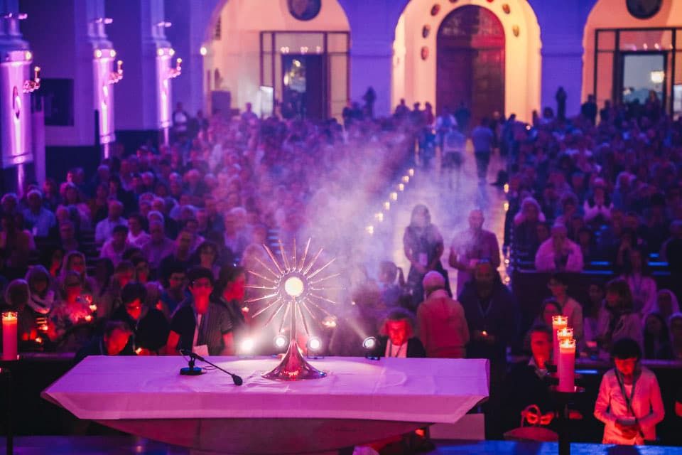 Eine beleuchtete Monstranz steht auf dem Altar. Die Kirche ist voll von Leuten, die das Allerheiligste anbeten. Das Licht ist warm und angenehem. Die Kirche ist stimmungsvoll beleuchtet