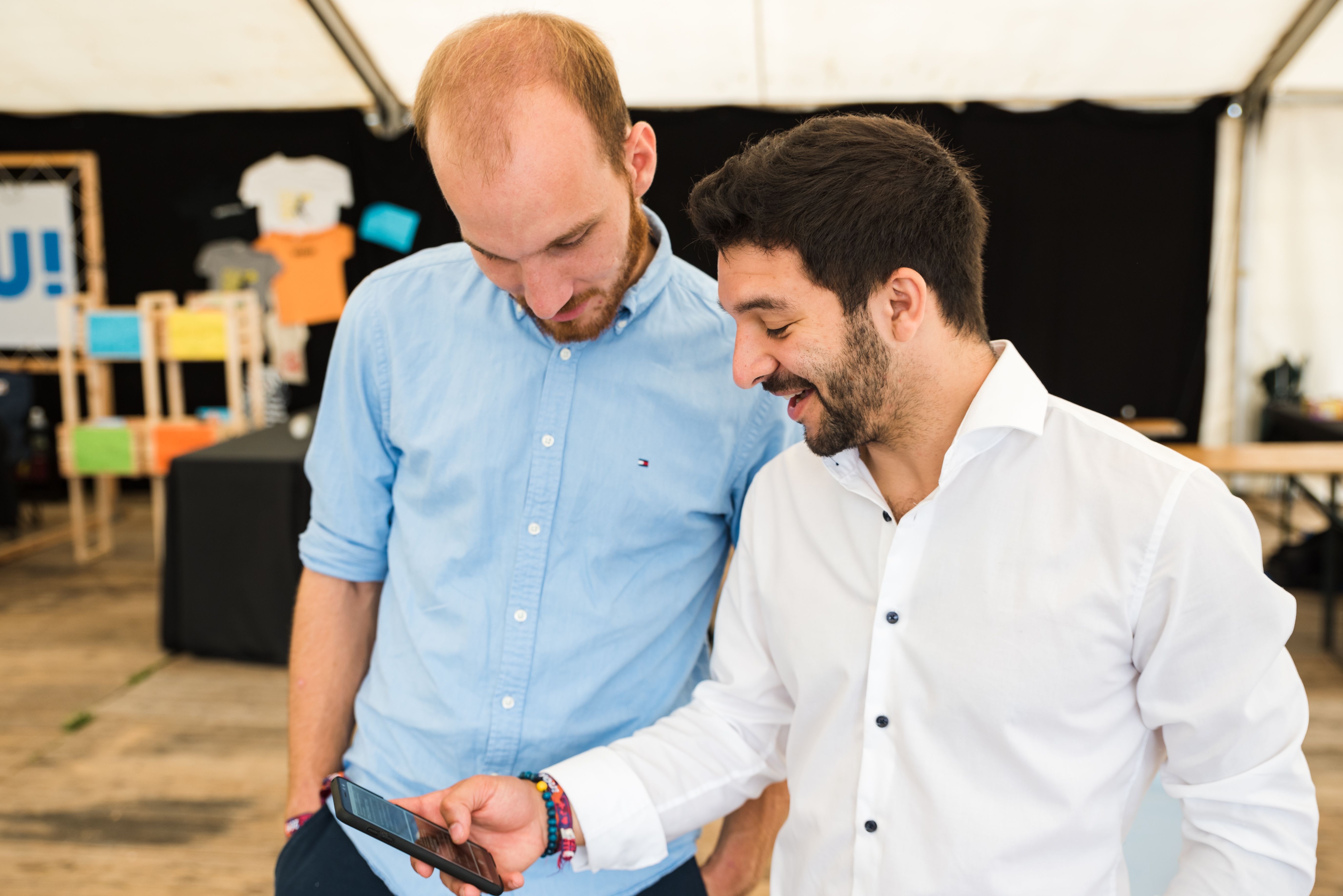 zwei Junge Männer schauen auf ihr Smartphone und strahlen vor Begeisterung. Im Hintergrund sieht man den Infostand eines katholischen Jugendmagazines