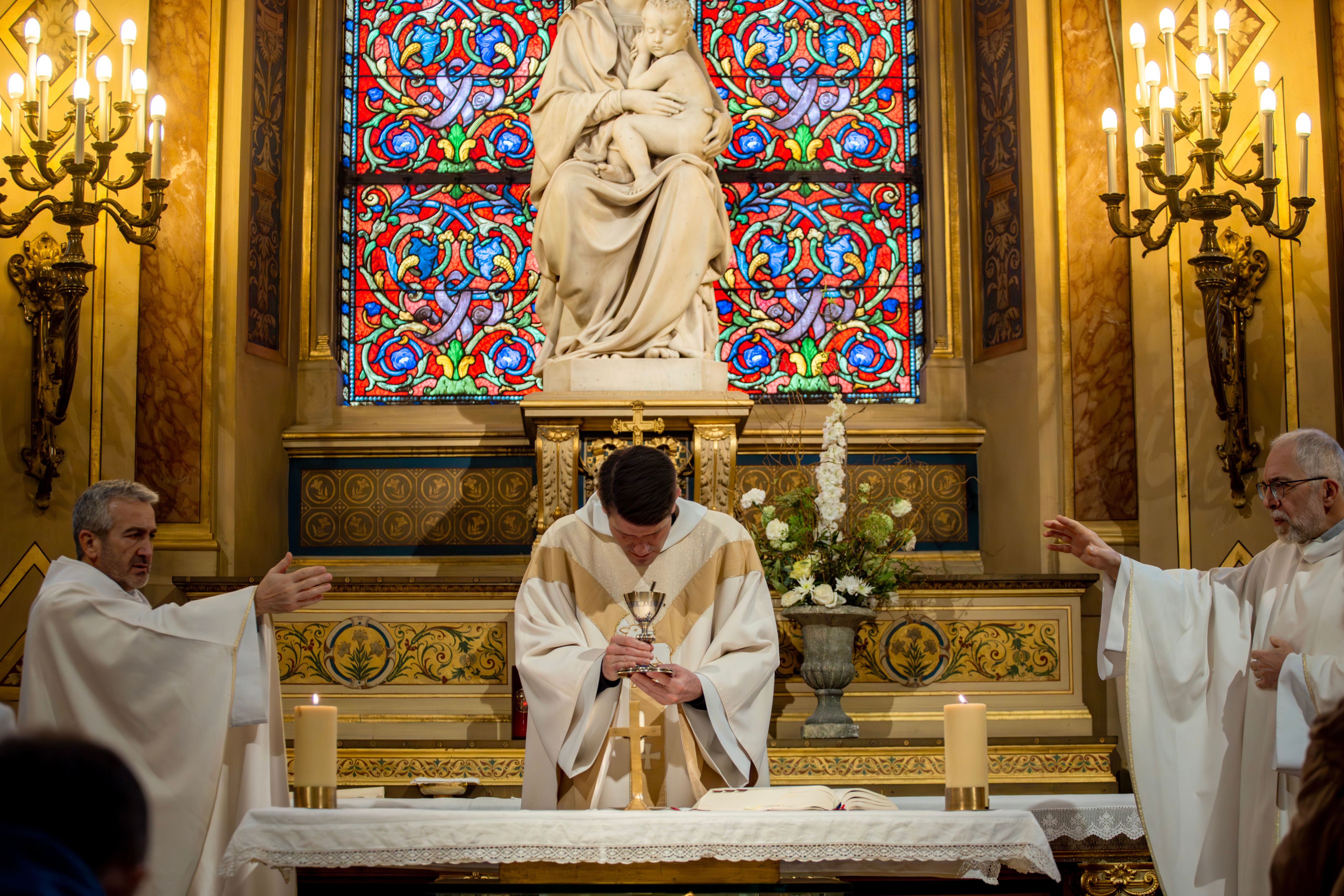 katholischer Gottesdienst in einer Kirche, drei Priester sprechen die Wandlungsworte, Priester in der Mitte verbeugt sich vor dem Kelch, im Hintergrund sieht man zwei Kerzenständer, eine Marienstatue und ein schillerndes Kirchenfenster