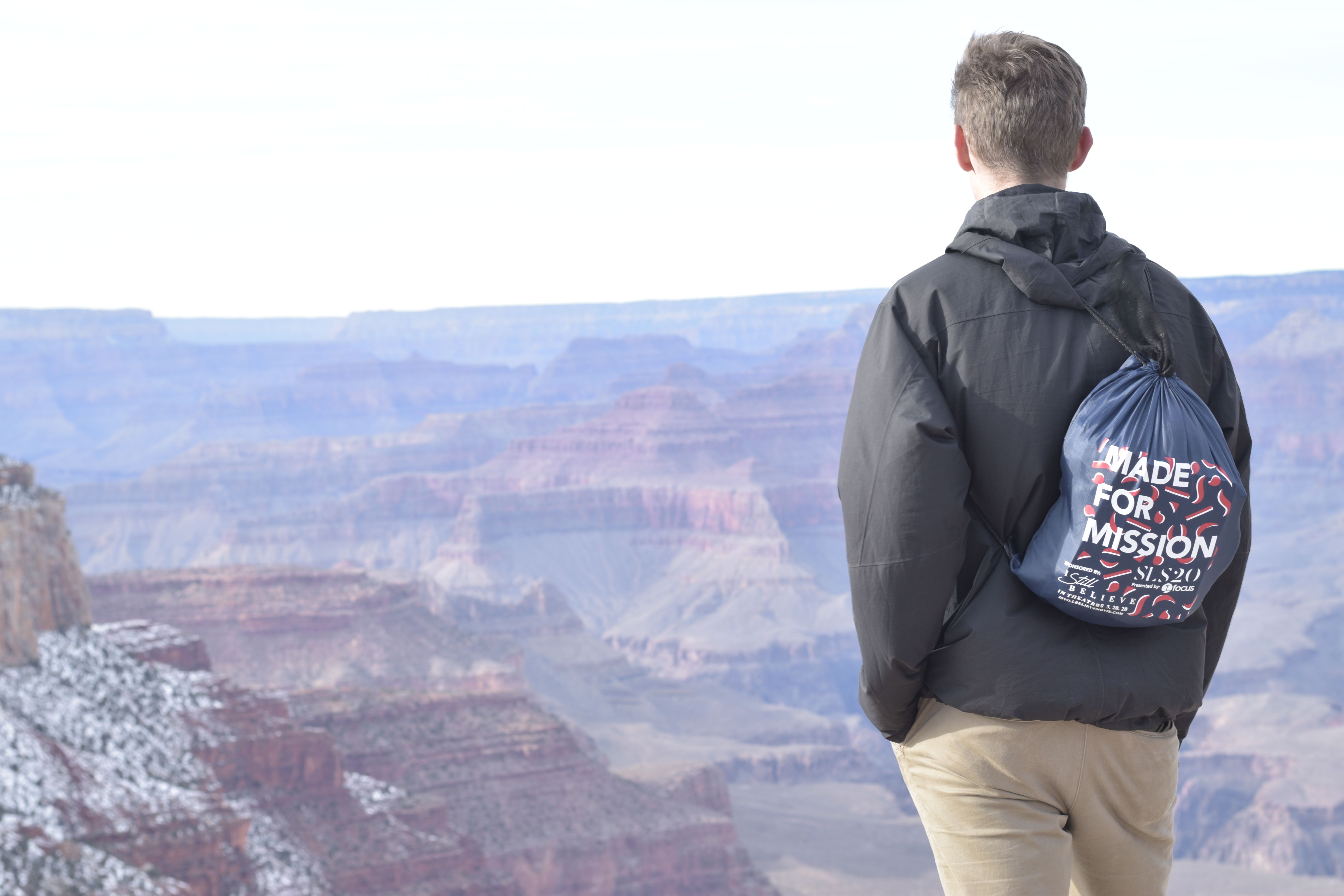 junger Student steht am Rande des Gran Canyon; Mann steht mit dem Rücken zur Kamera, er trägt einen Beutel der Seekconference am Rücken, auf dem "made for mission" steht