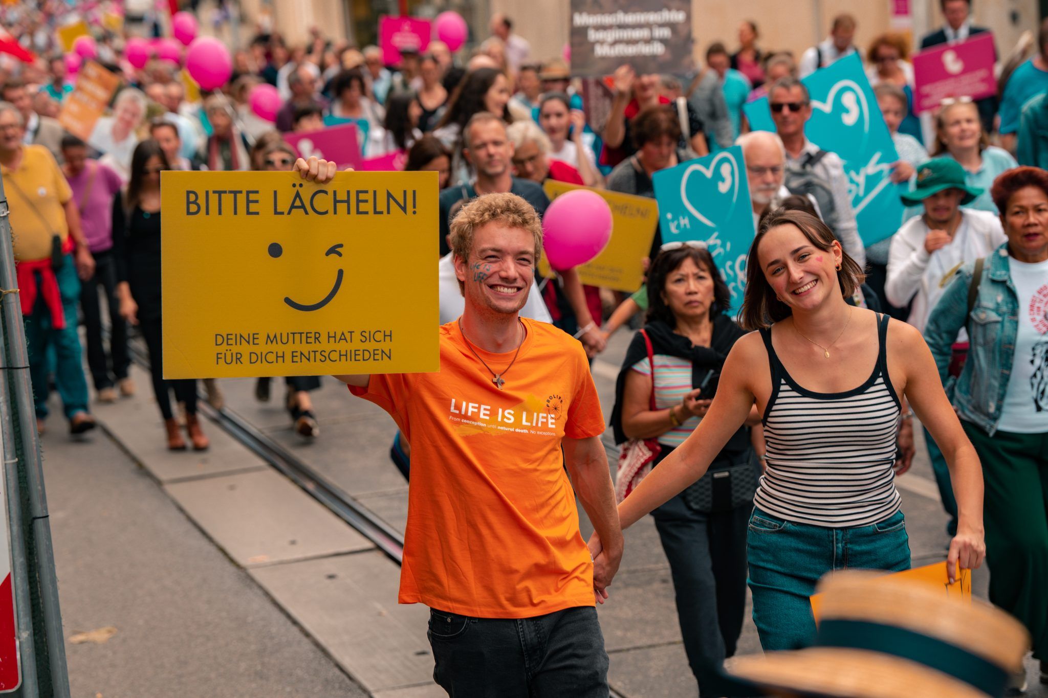 young man holds a sign saying "please smile", young woman walks laughing beside him, good atmosphere, joy of life becomes visible
