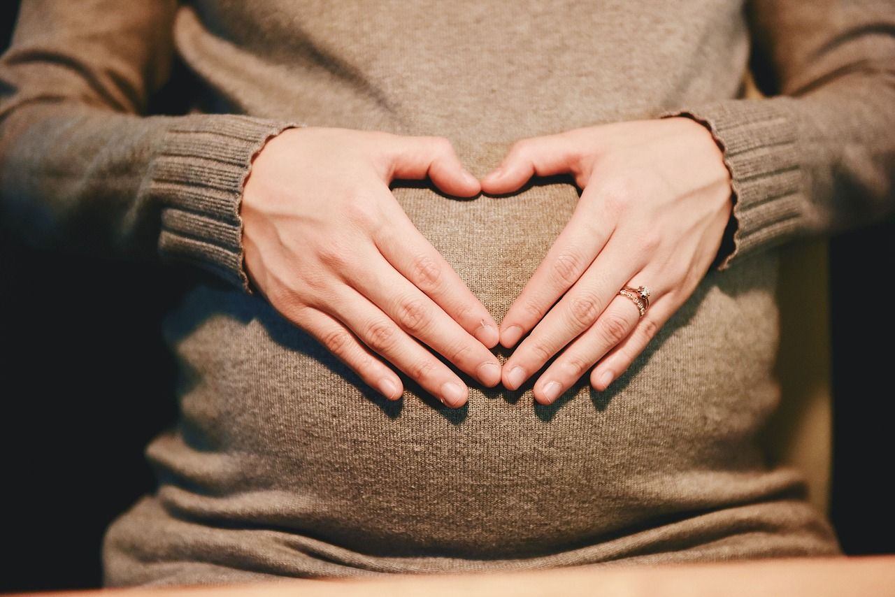 Schwangere Frau in braunem Wollkleid fasst mit Händen an ihren schwangeren Babybauch und formt ein Herz