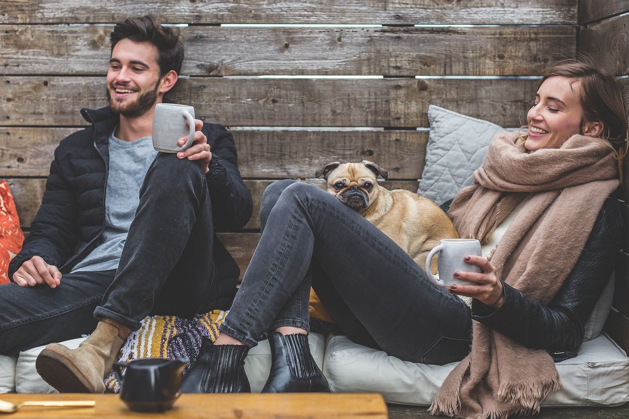 zwei Junge Leute sitzen bei Tasse Kaffee oder Tee beieinander und lachen. Ein Hund sitzt nebenbei. 