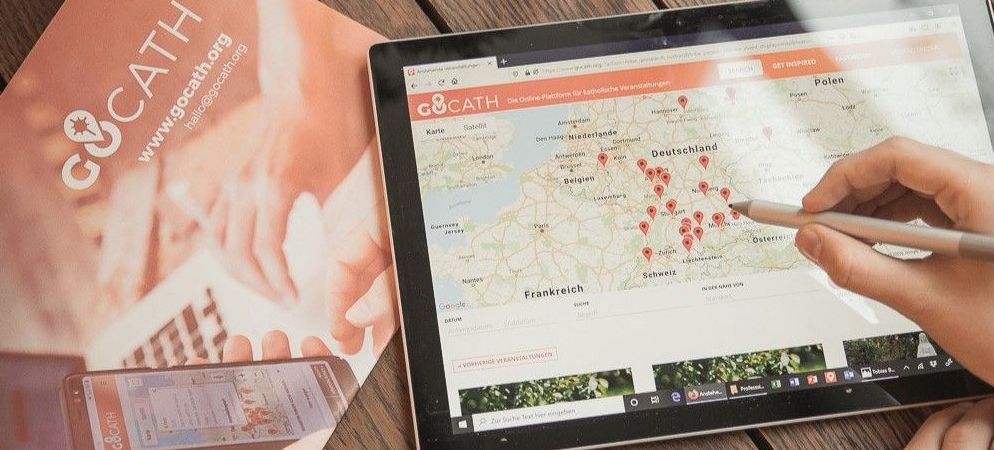 ein orangenes GOCATH-Plakat und ein Tablet mit der GOCATH-Veranstaltungsübersicht liegen auf einem hölzerenen Tisch. Ein User klickt mit einem Stift auf eine Veranstaltung