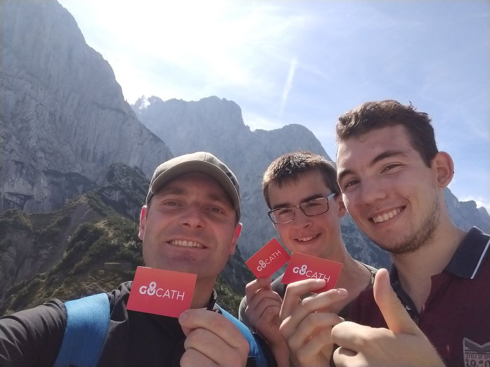 Priester und zwei junge Männer freuen sich, sind in den Bergen, die Sonne scheint, alle halten eine orangene GOCATH-Visitenkarte in der Hand
