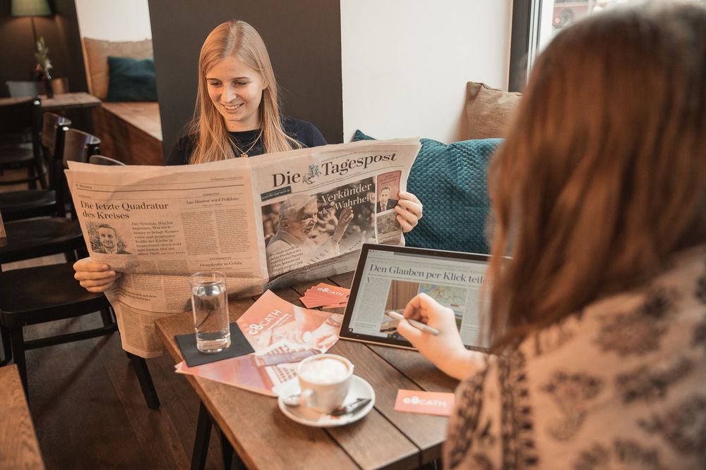 Presse, GoCATH in der Zeitung, Junge Frau liest in der Tagespost in einem Cafe. Mit ihr am Tisch sitzt eine Frau und zeigt mit Stift auf ihr Tablet