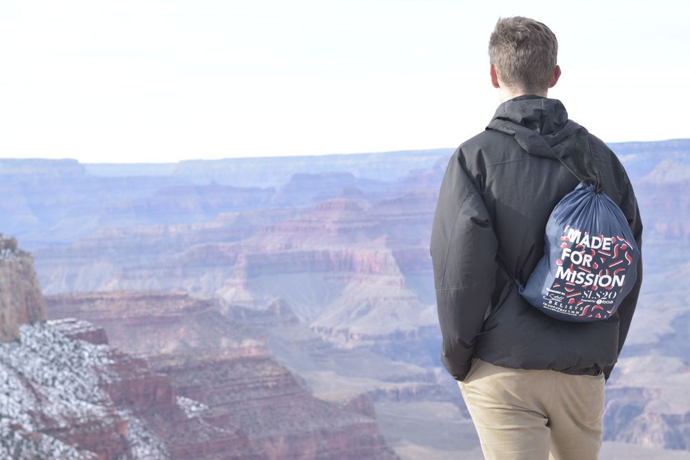 junger Student steht am Rande des Gran Canyon; Mann steht mit dem Rücken zur Kamera, er trägt einen Beutel der Seekconference am Rücken, auf dem "made for mission" steht
