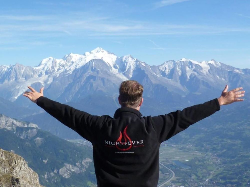 Mann mit ausgebreiteten Armen und Nightfever Jacke, im Hintergrund hohe Berge mit Schnee