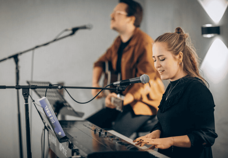 junge Frau spielt Klavier und singt modernen Lobpreis, ein Mann spielt nebendran Gitarre